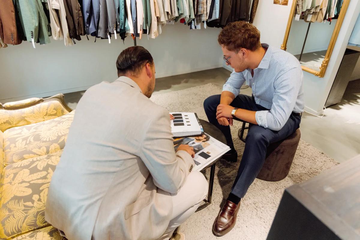 Conseiller en boutique montrant un nuancier de tissus à un client assis, essayage de costume en Belgique